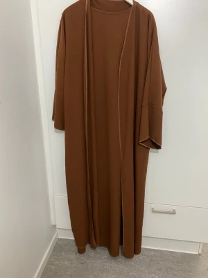 Brun lång kimono i bomull 2-delar - Säljer en stilren brun lång kimono 2 delar med vida ärmar och öppen front. Perfekt att slänga över en outfit för en avslappnad look. Tillverkad i mjuk bomull och har en enkel, minimalistisk design. Endast använd 2 gånger