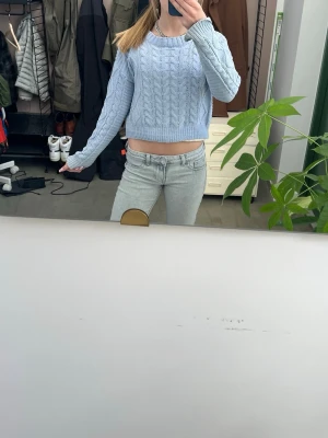 Ljusblå cropped stickad tröja - Säljer en ljusblå cropped stickad tröja med kabelstickat mönster. Tröjan har långa ärmar, rund halsringning och ribbade muddar vid ärmslut och nederkant. Perfekt för dig som gillar en avslappnad men trendig stil.