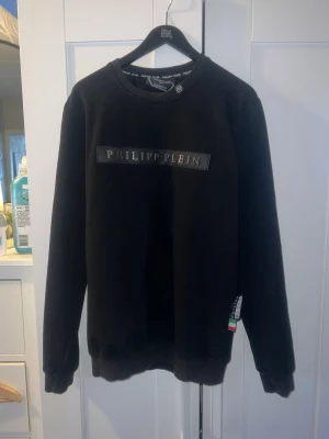 Svart sweatshirt från Philipp Plein - Snygg svart sweatshirt från Philipp Plein med logotyp i silver på bröstet. Tröjan har rund halsringning, ribbade muddar vid ärmslut och nederkant samt en liten detalj med italienska flaggan vid sidan. Perfekt för dig som gillar streetwear och exklusiva märken.