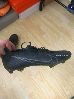 Nike Air Zoom svarta fotbollsskor - Snygga svarta Nike Air Zoom fotbollsskor med diskret Swoosh-logga i grått och gula detaljer på dobbarna. Skorna har snörning och en smal, sportig siluett i syntetmaterial som ger bra grepp på planen. Perfekta för dig som vill ha stilrena och snabba skor på matchen.