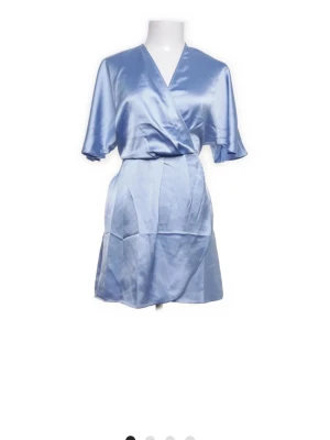 Ljusblå omlottklänning i satin - Snygg ljusblå kort omlottklänning i glansigt satinmaterial med v-ringning och korta ärmar. Perfekt för dig som vill ha en chic och trendig look med en touch av elegans.