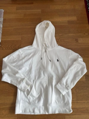Vit hoodie från Polo Ralph Lauren - Klassisk vit hoodie med dragkedja från Polo Ralph Lauren. Hoodien har huva med dragsko, två fickor framtill och den ikoniska lilla broderade loggan på bröstet. Perfekt för en clean och stilren look.