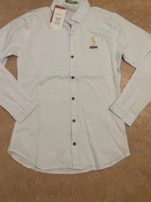 Vit skjorta från Polo Ralph Lauren - Klassisk vit skjorta från Polo Ralph Lauren med broderad logga på bröstet. Skjortan har lång ärm, knappar framtill och en stilren krage. Perfekt för en clean och tidlös look.