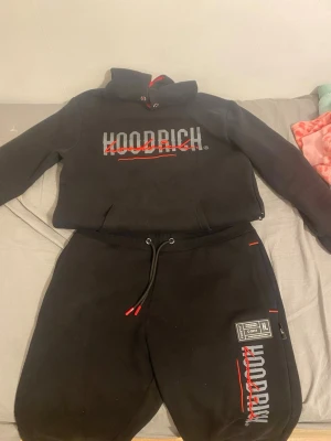 Svart Hoodrich loungewear set - Säljer ett svart loungewear set från Hoodrich med matchande hoodie och byxor. Setet har tryckt logga i vitt och rött på både tröja och byxa, samt röda detaljer på snören och huva. Perfekt för chill dagar hemma eller streetwear vibes.