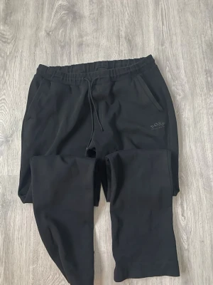 Hugo Boss byxor - Svarta mjukisbyxor från Hugo Boss med dragsko i midjan och två sidofickor. Byxorna har en stilren design med diskret BOSS-logga på ena benet. Perfekta för en chill och avslappnad stil.