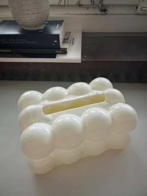 Vit bubblig servetthållare i plast - Säljer en unik servetthållare i vit plast med bubblig, modern design. Perfekt som inredningsdetalj på bordet eller i badrummet. Den har en rektangulär öppning upptill för enkel åtkomst till servetter.