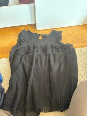 Svart blus från Zara Basic - Säljer en svart blus från Zara Basic med spetsdetaljer upptill och plisserad nederdel. Blusen är ärmlös och har en liten knapp i nacken. Perfekt för dig som gillar en chic och stilren look. Ganska liten för att vara xl, skulle säga kanske xs-m 