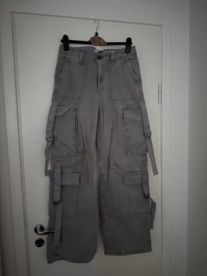 Grå cargobyxor med straps - Säljer ett par grå cargobyxor med bred passform och flera stora fickor på benen. Byxorna har coola straps och D-ringar längs sidorna för en edgy look. Tillverkade i ett slitstarkt bomullstyg, perfekta för dig som gillar streetwear och utilitystil.