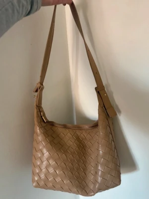 Beige flätad axelväska i läderimitation - Snygg beige axelväska med flätad design och justerbart axelband. Väskan har dragkedja upptill och är tillverkad i läderimitation. Perfekt för dig som gillar stilrena och trendiga accessoarer.