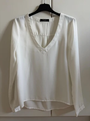 Vit blus med v-ringning från Vila - Stilren vit blus från Vila med djup v-ringning och långa ärmar. Blusen har en lös passform och är tillverkad i ett lätt, tunt tyg som ger ett snyggt fall. Perfekt att matcha med jeans eller kjol för en trendig look.