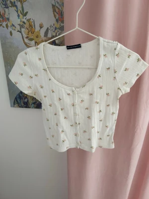 Vit blommig topp från Brandy Melville - Supersöt vit topp från Brandy Melville med små blommor, korta ärmar och knappar framtill.         🦢💓☁️