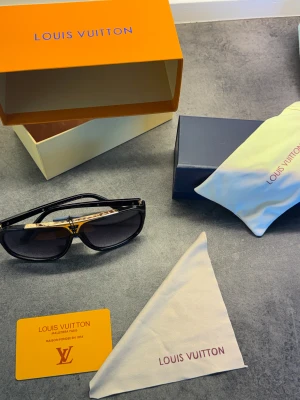 Louis Vuitton svarta solglasögon - Snygga svarta solglasögon från Louis Vuitton med gulddetaljer och klassisk logotyp på bågen. Glasögonen har en rektangulär form och levereras med originalförpackning, fodral och putsduk. Perfekt accessoar för att lyfta din stil.
