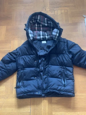 Svart pufferjacka från Burberry - Snygg svart pufferjacka från Burberry med klassiskt rutigt foder i huvan. Jackan har dragkedja och tryckknappar framtill, två fickor med dragkedja och en huva.  går även att ta av ärmarna och göra den till en väst 