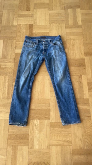 Blå raka jeans från Nudie Jeans - Säljer ett par klassiska blå jeans från Nudie Jeans med raka ben och femficksmodell. Jeansen har snygga slitningar och kontrastsömmar samt Nudie-logga på bakfickorna. Perfekta för en avslappnad och trendig stil.