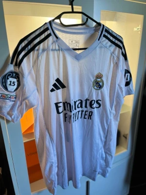 Real Madrid Mbappé matchtröja - Vit Real Madrid fotbollströja från Adidas med svarta detaljer och tryck. Tröjan har klubbmärke, sponsorlogga och nummer 9 med Mbappé på ryggen. Champions League-märke på ärmen och klassisk V-ringning. Perfekt för fotbollsfans!