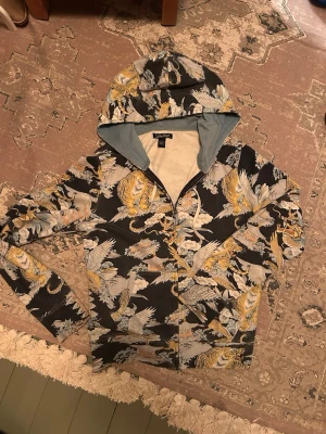 Vintage zipup Lucky Brand -  Lucky Brand holdie med dragkedja och huva. Hoodien har ett detaljerat mönster med drakar och blommor i gult, vitt och blått. Tillverkad i 100% bomull och har ljusblått innerfoder i huvan. Storlek M men passar bra också S.