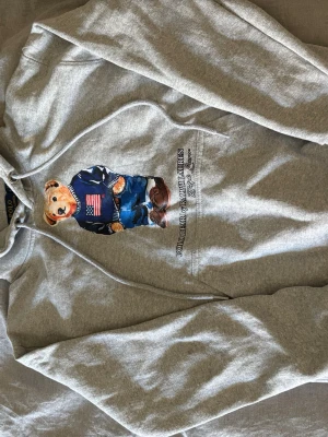 Grå hoodie Polo Ralph Lauren björn - Snygg grå hoodie från Polo Ralph Lauren med ikonisk björn på bröstet som bär en blå tröja med amerikansk flagga. Klassisk huva med dragsko och känguruficka framtill. Perfekt för en avslappnad och trendig stil. Använd en gång! Säljer nu för att den för liten! 