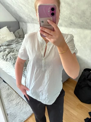 Vit-blå randig blus  - Säljer en vit-blå randig blus med korta ärmar och ståkrage. Blusen har fina veck framtill och knappar vid halsen. Tillverkad i ett lätt och luftigt tyg som ger en avslappnad men ändå stilren look. Den är i storlek 40 men väldigt liten i storleken, passar mer som en S 