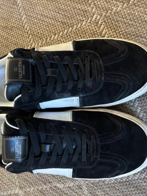 Flycrews  - Snygga sneakers från Valentino i svart mocka med vita läderdetaljer på sidorna. Klassisk låg modell med svarta skosnören och vit sula. Märkeslogga på plösen och stilren design som passar till många outfits. Alla nitar är kvar! Boxen ingår även.