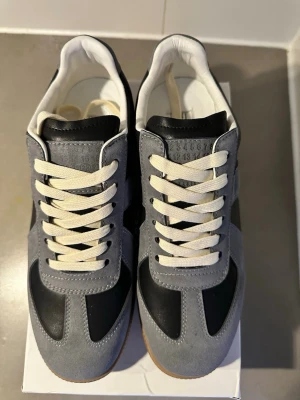 Grå och svarta sneakers Maison Margiela kvitto finns  - Maison Margiela sneakers med grå mockadetaljer, svarta paneler i läder och vita snören. Klassisk siluett med låg profil och gummisula i brunt. Ikonisk design med diskret präglad logga på plösen och vit insida.