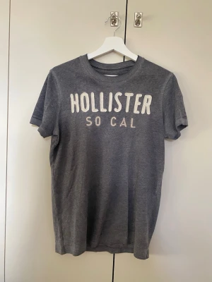 Grå t-shirt från Hollister - Snygg grå t-shirt från Hollister med vit text 'HOLLISTER SO CAL' på bröstet. Klassisk rund halsringning och korta ärmar. Perfekt för en avslappnad och stilren look. Tillverkad i mjuk bomull för extra komfort.
