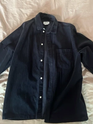 Svart manchesterskjorta från Weekday - Snygg svart skjorta från Weekday i manchestertyg. Klassisk krage, vita knappar och bröstficka. Perfekt för dig som gillar en stilren och enkel look. Skjortan har lång ärm och rak passform.