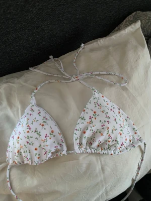 Vit blommig trekantsbikini - Söt trekantsbikini i vitt med små blommiga mönster i rosa, orange och grönt. Bikinin har tunna knytband i nacken och ryggen för justerbar passform. Perfekt för soliga dagar på stranden.