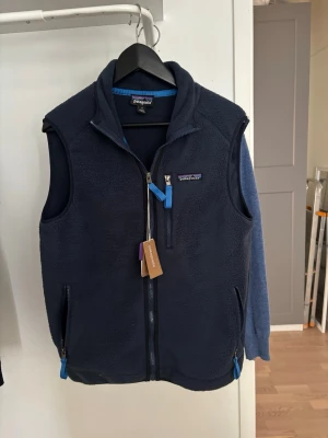 Mörkblå fleeceväst från Patagonia - Mörkblå fleeceväst från Patagonia med dragkedja framtill, två sidofickor och en bröstficka med blå detaljer. Västen har hög krage och klassisk passform, perfekt för lager-på-lager. Materialet är mjuk fleece som håller dig varm under kyliga dagar.