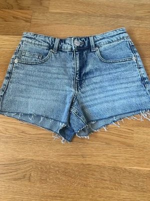Blå jeansshorts med fransig kant VMD - Snygga blå jeansshorts från Vero Moda med medel midja och fransig kant nertill. Klassisk femficksmodell med knapp och dragkedja framtill. Perfekta för sommaren och lätta att matcha med allt.