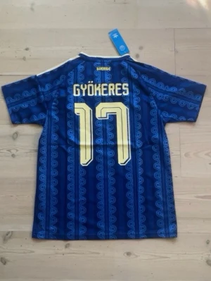 Sverige fotbollströja Gyökeres 2026 - Snygg blå fotbollströja från Adidas med Sveriges landslagsemblem och Gyökeres nummer 17 i gult. Säljer denna tröja för att den var för liten, helt helt ny.