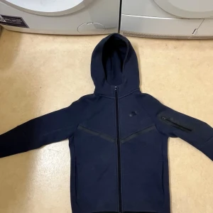 Mörkblå Nike hoodie med dragkedja - Barn storlek Snygg mörkblå hoodie från Nike med hel dragkedja framtill och huva. Har en diskret logga på bröstet och en praktisk ficka med dragkedja på ena ärmen. Perfekt för en sportig och avslappnad stil.