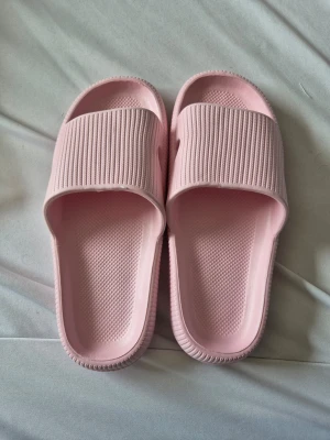 Ljusrosa ribbade tofflor i gummi - Säljer ett par ljusrosa tofflor med ribbad ovandel och räfflad sula. Tillverkade i mjukt och flexibelt gummi för extra komfort. Perfekta att glida i hemma eller på stranden. Snygg minimalistisk design med öppen tå och struktur på fotbädden.