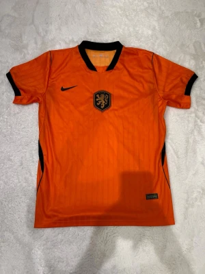 Holland Nike fotbollströja orange - Snygg orange fotbollströja från Nike med svarta detaljer och KNVB-emblem på bröstet. Tröjan har korta ärmar, svart krage och är tillverkad i ett lätt och ventilerande material som passar perfekt för träning eller match.