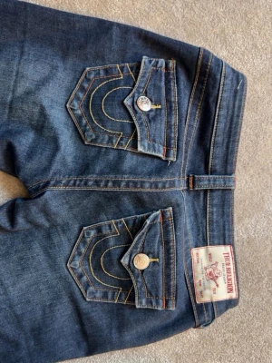 True Religion mörkblå bootcut jeans - kolla sista bilden, Slitningar från att ja råkat gå på dom därav priset. Säljer ett par mörkblå bootcut jeans från True Religion med ikoniska kontrastsömmar och stora fickor bak med lock och metallknappar. Klassisk femficksmodell i stretchigt denimtyg som sitter snyggt och ger en retrovibe.