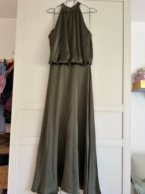 Långklänning - Snygg olivgrön långklänning med halterneck och lös passform upptill. Klänningen har ett elegant fall och är tillverkad i ett glansigt material som ger en lyxig känsla. Perfekt för dig som vill ha en stilren och trendig look. Aldrig använd med lapp kvar. 