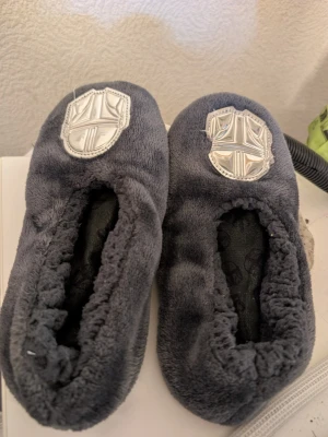 Mörkgrå Star Wars tofflor stl 34-35 - Mysiga mörkgrå tofflor i mjuk fleece med Star Wars-motiv på ovansidan och halkskydd med Mandalorian-symboler under sulan. Perfekta för att chilla hemma och hålla fötterna varma.fint skick dock har en liten bit av silver märker lossat annars jätte fina 
