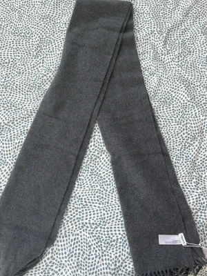 Grå halsduk från Filippa K i ull - Stilren grå halsduk från Filippa K i mjuk ull. Klassisk rektangulär form med diskreta fransar i ena änden. Perfekt för att hålla värmen och ge en clean look till din outfit.