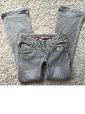 Grå jeans från Rainbow Country - Snygga grå jeans från Rainbow Country med utsvängda ben. Klassisk femficksmodell med dekorativt märke bak och inslag av blommigt tyg i ena fickan. Perfekta för en avslappnad och trendig look.