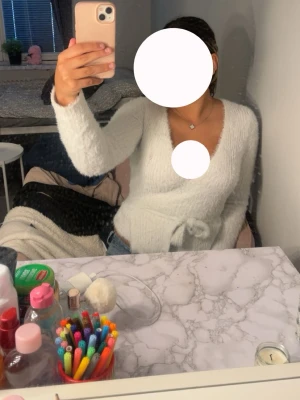 Vit fluffig omlottkofta - Mysig vit omlottkofta med knytband i midjan och lång ärm.  Passar snyggt till jeans eller kjol. Har ett litet hål vid bröstet men syns inte 