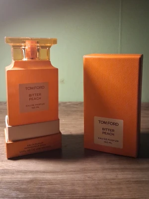 Tom Ford Bitter Peach Eau de Parfum - Tom Ford Bitter Peach Eau de Parfum 100 ml med en snygg orange glasflaska och matchande kartong. Flaskan har en fyrkantig form och ett elegant lock i transparent orange plast. Doften är lyxig och fruktig, perfekt för dig som gillar unika parfymer.
