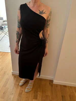 Svart oneshoulder klänning med slits - Snygg svart oneshoulder klänning med cut-out och snörning i sidan samt hög slits framtill. Klänningen har en kroppsnära passform och är tillverkad i ett mjukt, stretchigt material som sitter skönt på kroppen. Perfekt för dig som vill sticka ut.