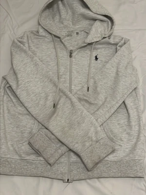 Ljusgrå hoodie från Polo Ralph Lauren - Ljusgrå zip-hoodie från Polo Ralph Lauren med dragsko i huvan och klassisk broderad logga på bröstet. Mjuk bomullsblandning, fickor framtill och ribbade muddar. Perfekt för en chill och stilren look.