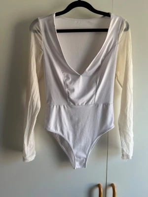 Vit långärmad body med djup V-ringning - Snygg vit body med långa ärmar och djup V-ringning framtill. Tillverkad i ett stretchigt och mjukt material som sitter tight mot kroppen. Perfekt att styla med jeans eller kjol för en trendig look. Endast provad katt finns i hem!