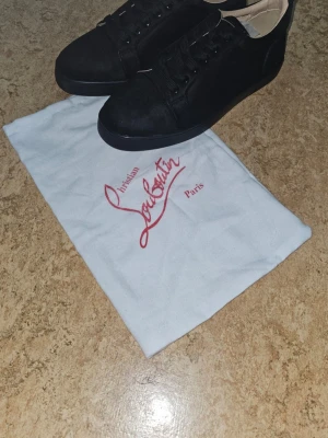 Svarta Christian Louboutin sneakers - Snygga svarta sneakers från Christian Louboutin med klassisk röd sula och diskret logga på sidan. Skorna har lågt skaft, svarta skosnören och är tillverkade i läder. Perfekta för dig som vill sticka ut med exklusiva detaljer och stilren design. Storlek 44 finns i 43