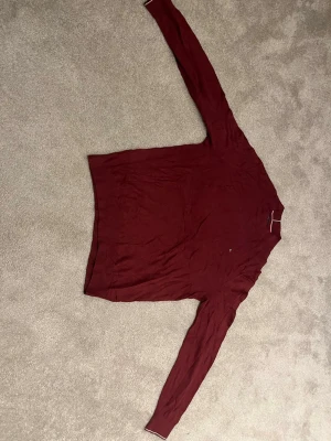Vinröd stickad tröja Tommy Hilfiger - Snygg vinröd stickad tröja från Tommy Hilfiger i mjuk pima bomull och kashmir. Tröjan har långa ärmar och klassisk rund halsringning med diskreta Hilfiger-detaljer i kragen. Perfekt för dig som gillar stilrena och bekväma plagg.