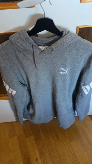 Grå hoodie från Puma med tryck - Snygg grå hoodie från Puma med vit logga broderad på bröstet och tryck på ärmarna. Hoodien har snörning i huvan och är tillverkad i mjuk bomullsblandning. Perfekt för en avslappnad och sportig stil.