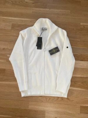 Vit stickad ziptröja Stone Island - Vit stickad ziptröja från Stone Island med hög krage och ribbad struktur. Tröjan har hel dragkedja framtill och klassisk logotyp på ärmen. Perfekt för lager-på-lager och stilren look.