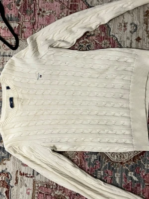 Vit kabelstickad tröja från Gant - Kabelstickad vit tröja från Gant med rund halsringning och diskret logga broderad på bröstet. Tröjan har långa ärmar och ribbade muddar vid ärmslut och nederkant. Perfekt för en klassisk och stilren look.