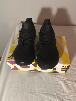 Svarta Adidas Ultraboost sneakers - Snygga helsvarta Adidas Ultraboost sneakers med stickad ovandel och klassiska tre ränder på sidan. Skorna har en chunky Boost-sula för maxad komfort och sportig look. Perfekta för dig som gillar streetstyle och vill ha en clean och modern sneaker.