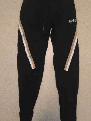 Svarta mjukisbyxor från BOSS - Svarta mjukisbyxor från BOSS med vita och beige ränder längs benen. Byxorna har dragsko i midjan och smal passform med muddar vid bensluten. Perfekta för en avslappnad och trendig stil.